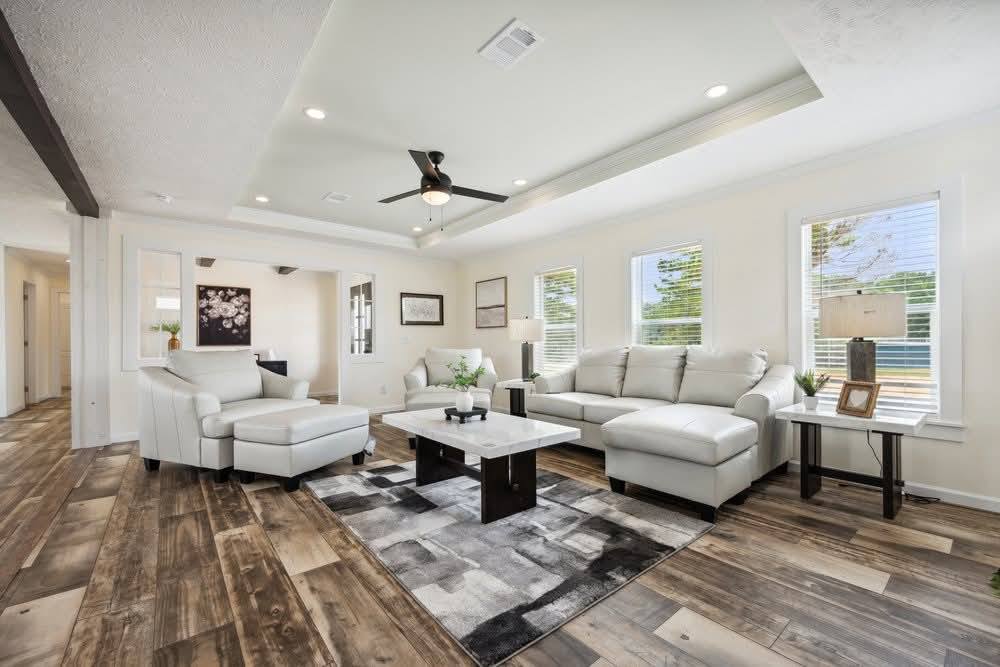 Spacious living room with white sofas and an armchair on a patterned rug. Wooden floor, large windows, ceiling fan, and soft lighting create a cozy ambiance.