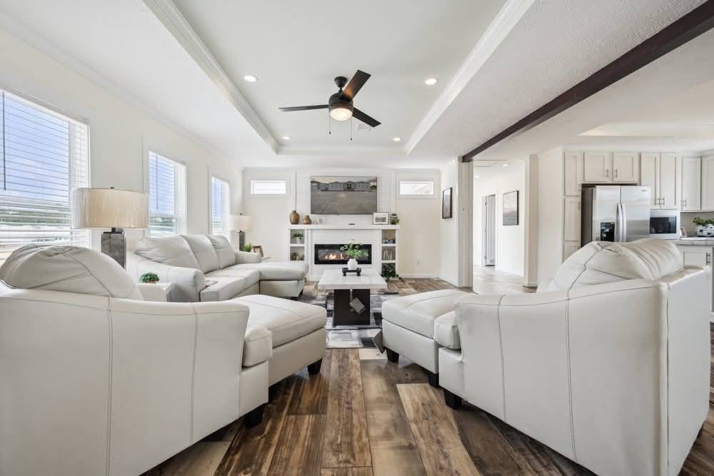 Bright, modern living room with white sofas, wood floor, and ceiling fan. Fireplace and TV centered on back wall, kitchen visible on right.