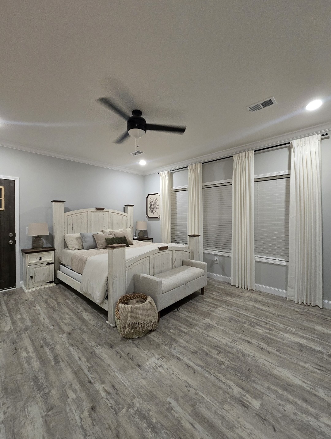 Spacious bedroom with a rustic bed, light wood flooring, soft gray walls, and a ceiling fan. Large windows with white curtains add an airy feel.