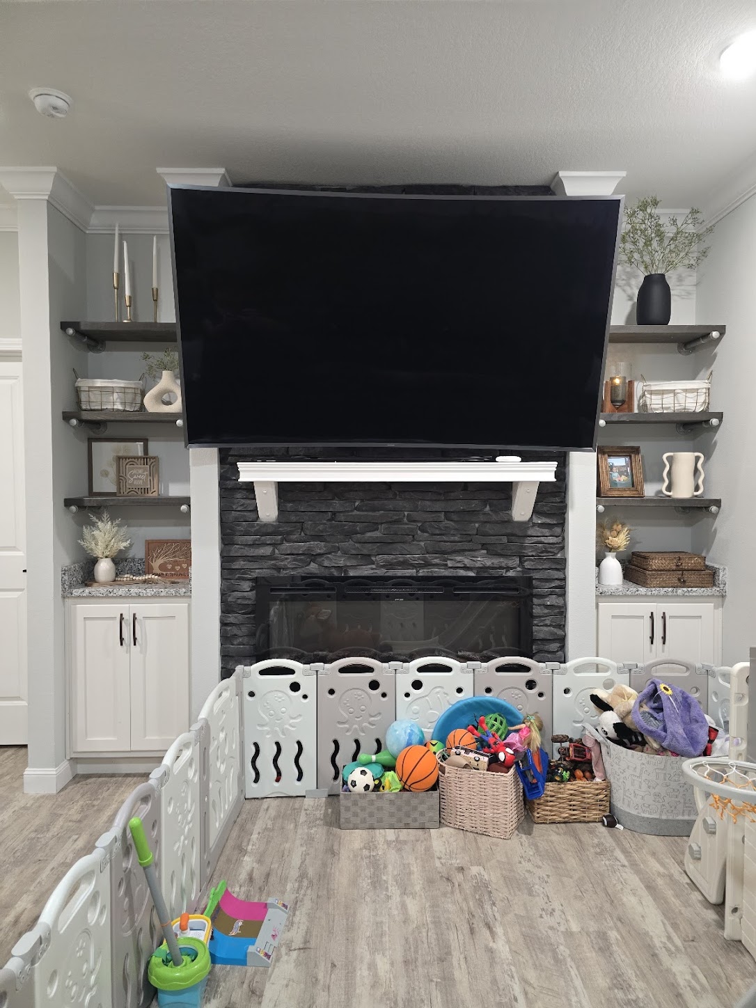 A living room features a large TV above a stone fireplace, flanked by decorative shelves with vases and photos. A playpen filled with toys adds a playful touch.
