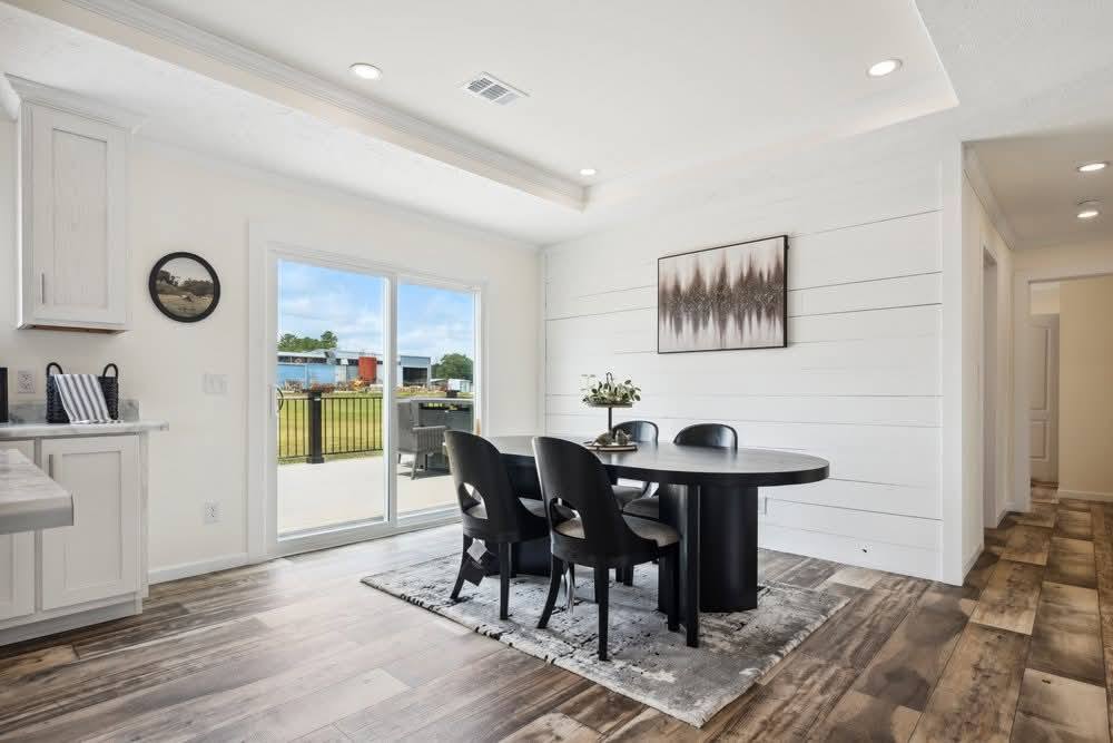 Bright dining area with a round black table, four chairs, and a potted plant centerpiece. Light wood floors and a large window showing an outdoor view.