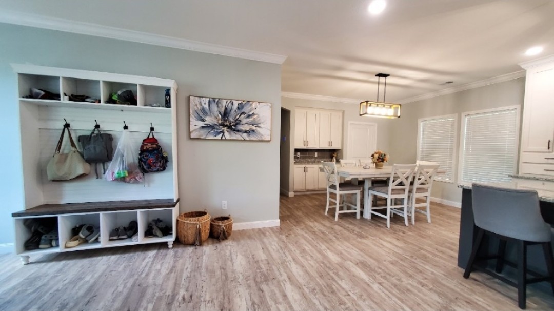 Spacious modern foyer and dining area with light wood flooring. Features a bench with hooks and storage, a large floral painting, and a dining table for six. Bright and inviting atmosphere.