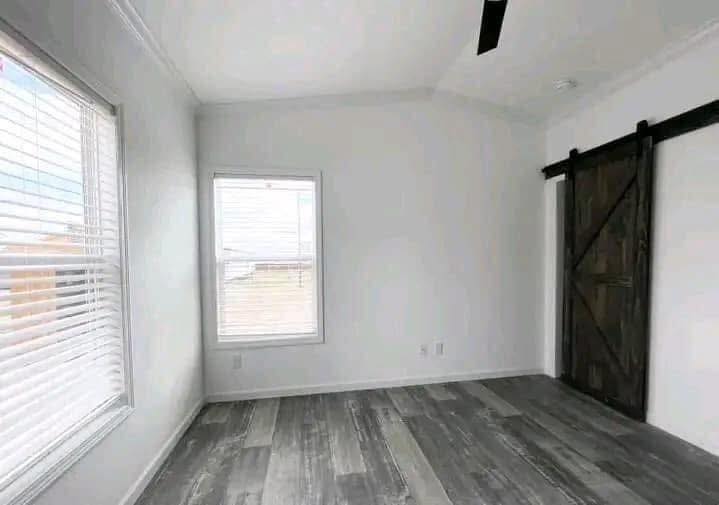 A minimalist empty room with gray wood flooring, white walls, and a rustic wooden sliding barn door. Two windows let in soft natural light.