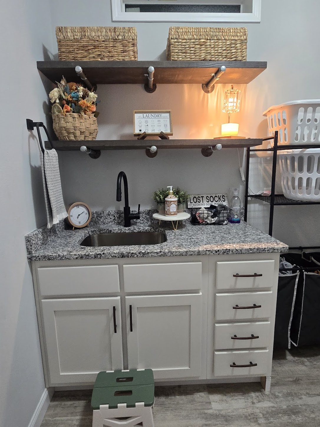 Laundry room with granite countertop, white cabinets, black faucet, and `Lost Socks` sign. Shelves hold baskets and a warm lamp, creating a cozy feel.