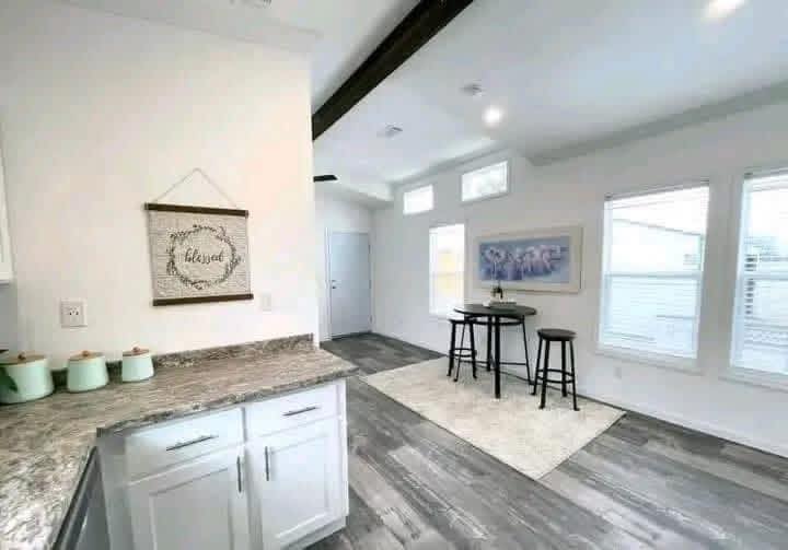 Modern kitchen and dining area with light wood flooring. White cabinets, green canisters, and a "blessed" sign adorn the kitchen. A round table with stools sits on a rug by large windows, creating a bright, welcoming space.