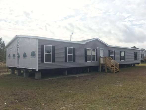 A gray two-section mobile home on concrete blocks with white trim and a small wooden porch, set on a grass field under a partly cloudy sky.