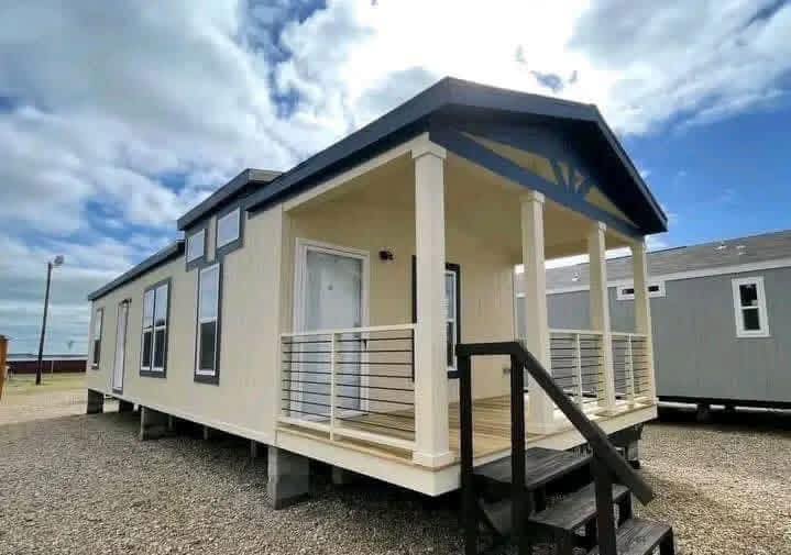 Modular home with cream siding and dark trim, featuring large windows and a covered porch. Set on gravel under a partly cloudy sky, conveying a modern, minimalist feel.