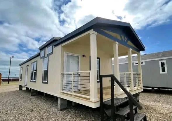 A beige modular home with a small front porch, railings, and steps is set on a gravel lot under a partly cloudy sky, conveying a sense of simplicity and modernity.