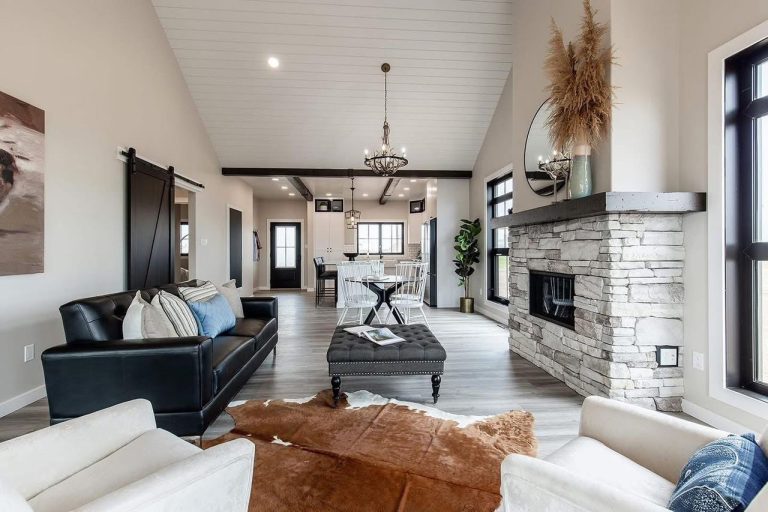 Spacious living area with high ceiling features a stone fireplace, black leather sofa, light chairs, cowhide rug, and a dining area with chandeliers.
