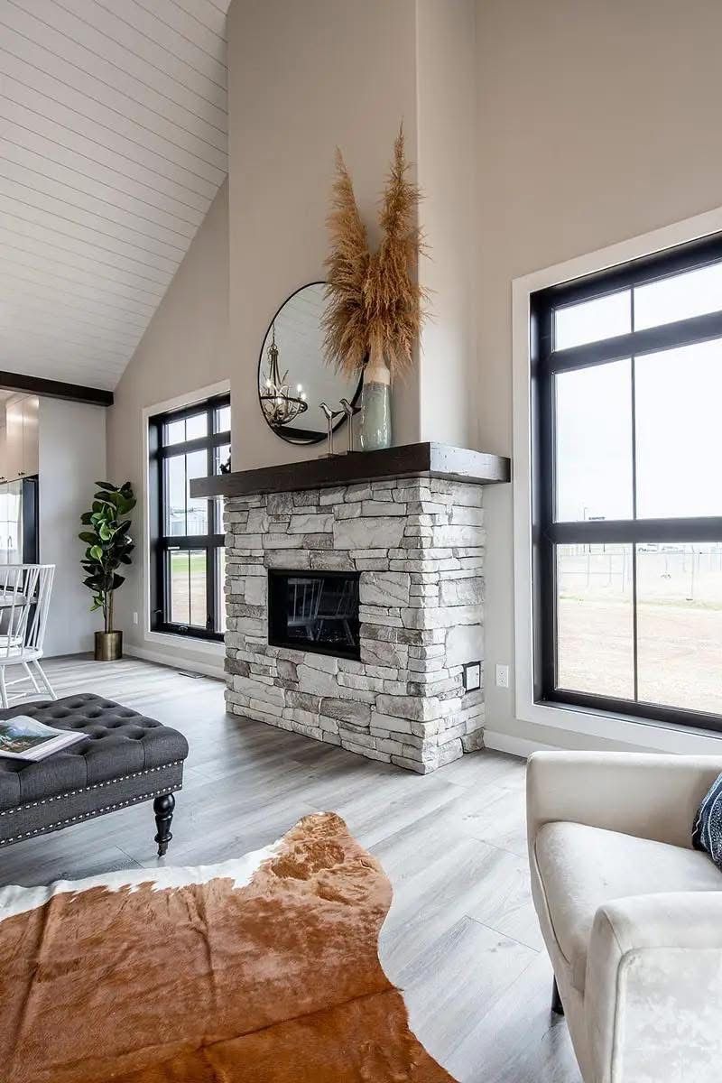 Modern living room with a stone fireplace and a round mirror above. Natural light floods through large windows, highlighting pampas grass decor and a cozy cowhide rug.