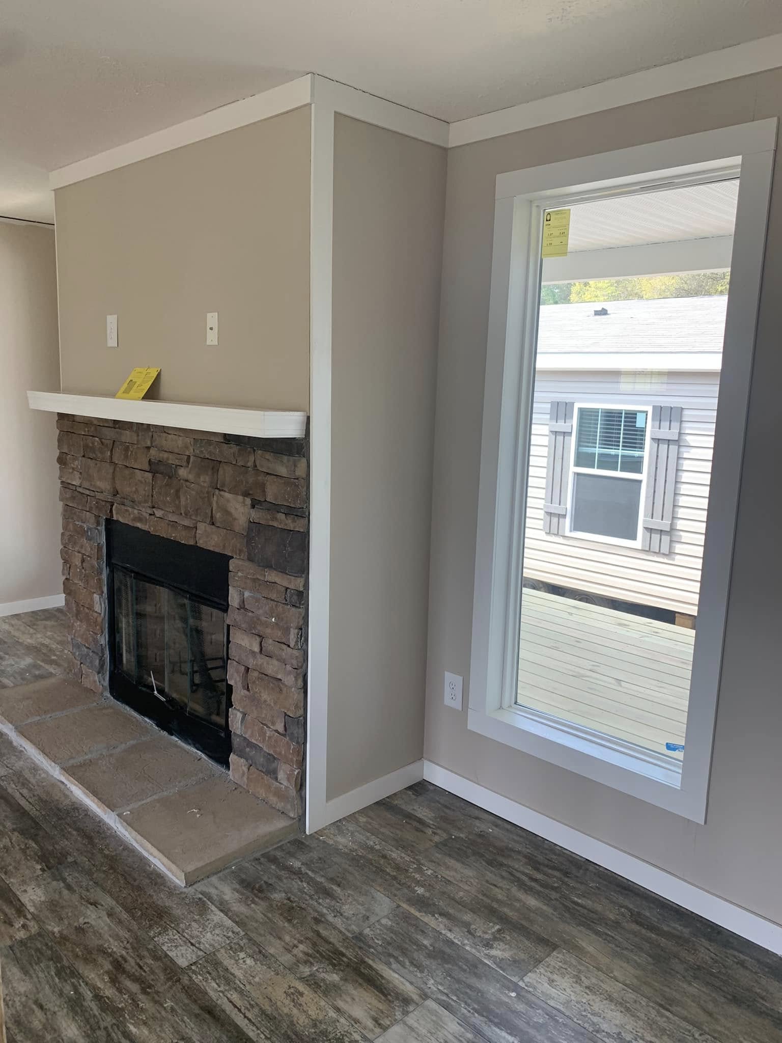 Living room with a stone fireplace, wooden floor, and a large window showing an outdoor view of another house. Warm and inviting atmosphere.