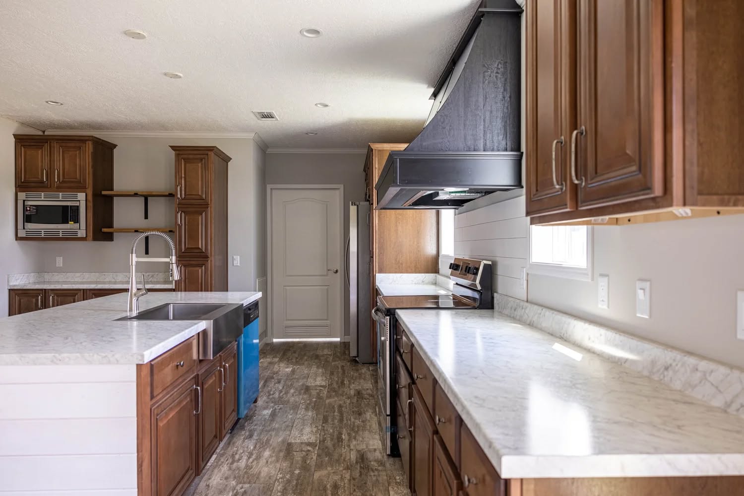 A modern kitchen with wooden cabinets and light marble countertops. It features an island with a metal sink, stainless steel appliances, and a range hood. The room is bright and spacious with natural light from a side window.
