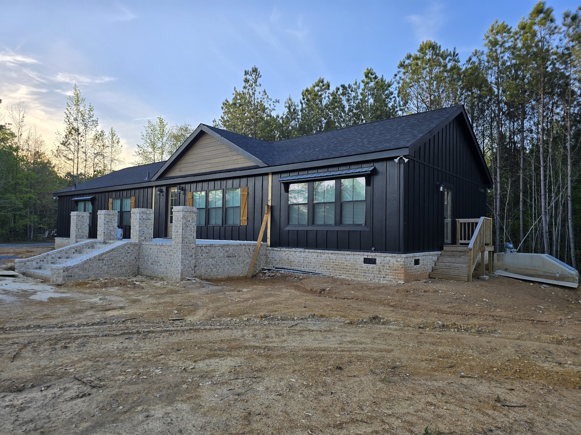 Single-story black house with brick foundation amid a wooded area. It has multiple windows, two staircases, and a clear blue sky overhead.