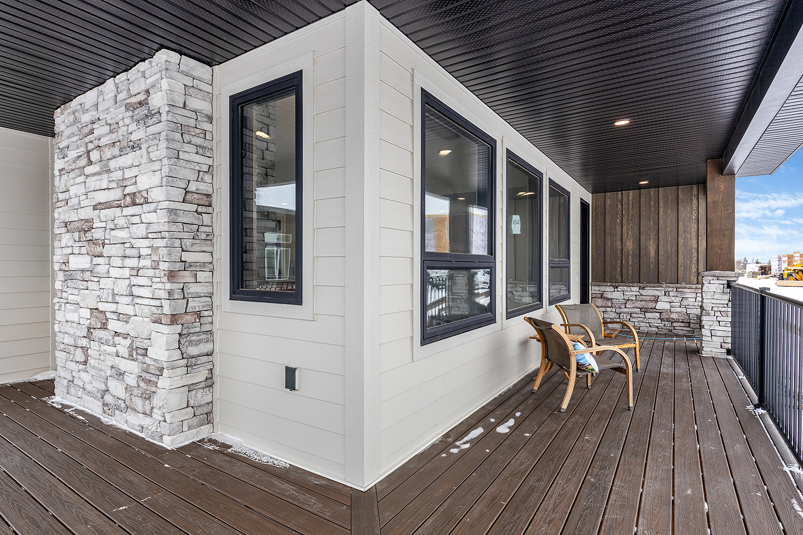Covered balcony with stone accent wall, dark wood ceiling, and wooden deck. Two wooden chairs face the railing under a bright, partly cloudy sky.