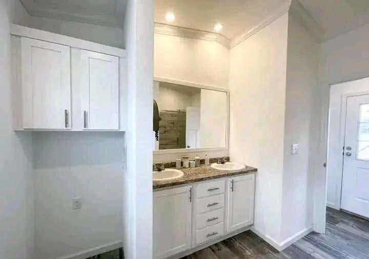 A bright, modern bathroom with dual sinks on a granite countertop, white cabinets, a large mirror, and wood-style flooring, conveying a clean and elegant feel.