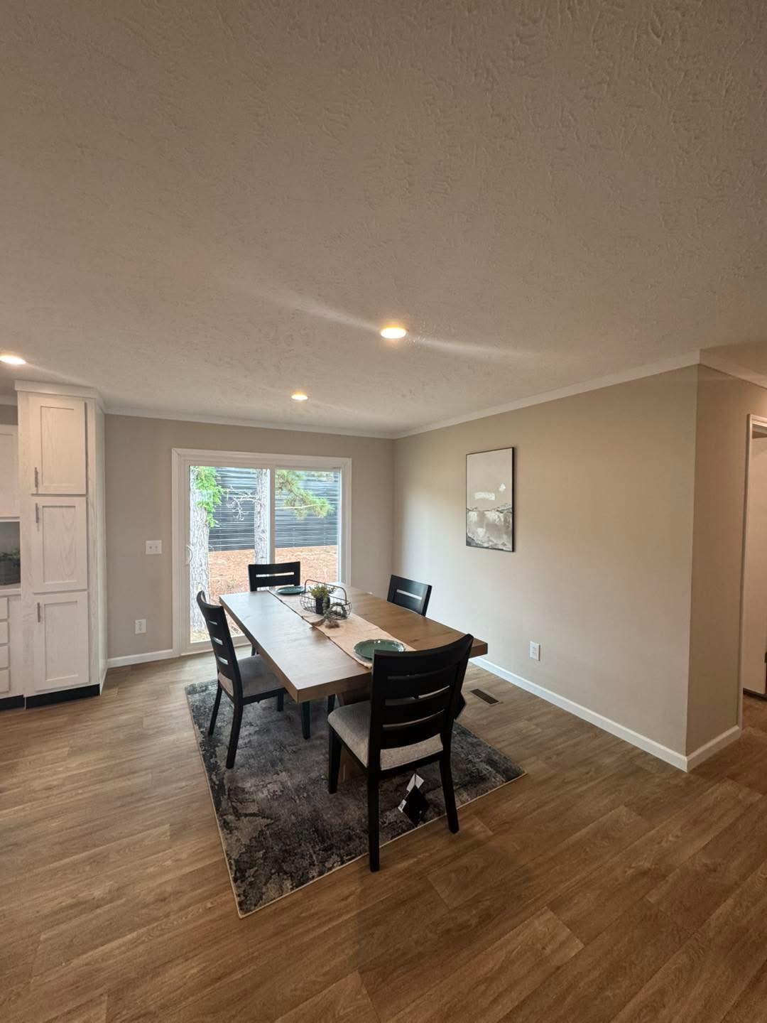 Modern dining room with wooden table and four black chairs on a textured rug. Bright natural light streams from a large window, creating an inviting atmosphere.