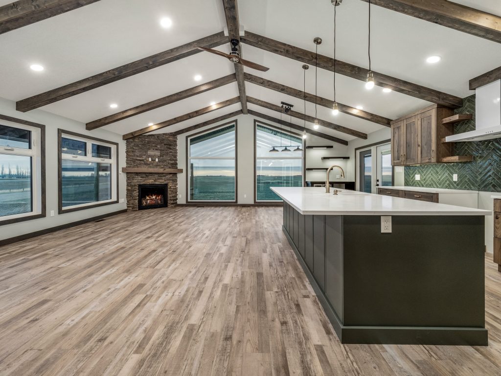 Spacious modern kitchen with wooden ceiling beams, large windows showing sunset, stone fireplace, and long island counter with pendant lights. Warm, inviting tone.