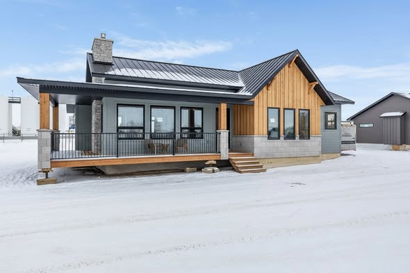 Modern house with wooden and stone accents on a snowy landscape. Features a metal roof, chimney, and a porch. Bright, clear winter sky.