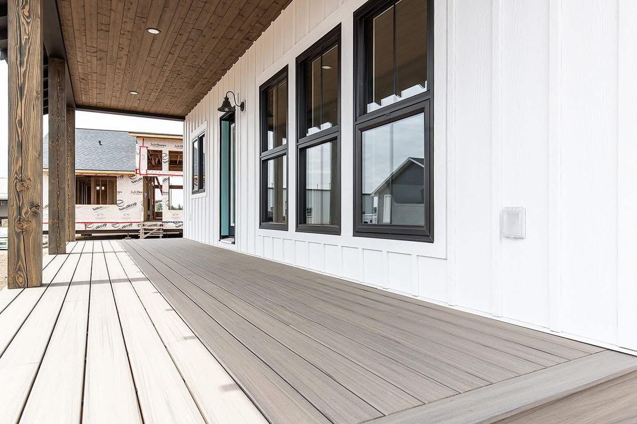 A modern porch with gray wooden decking and a white paneled wall featuring large black-framed windows. The porch has a wooden ceiling and support beams.