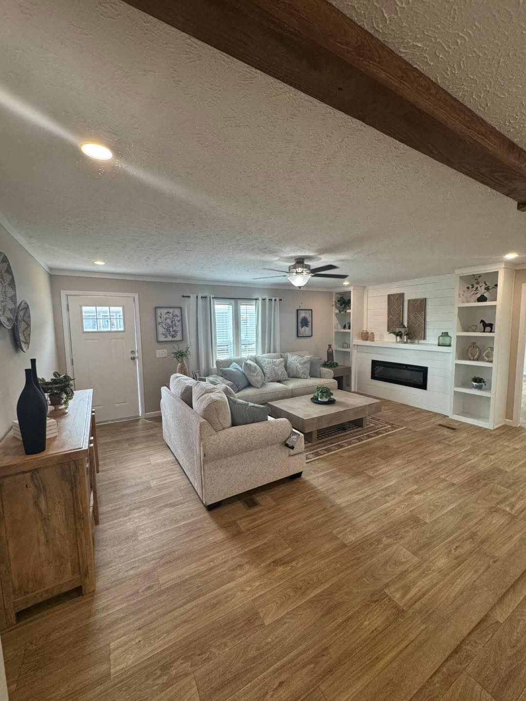 Spacious living room with light wood floors, beige sectional sofa, modern fireplace, and decorative shelves. Bright, airy atmosphere with natural light.