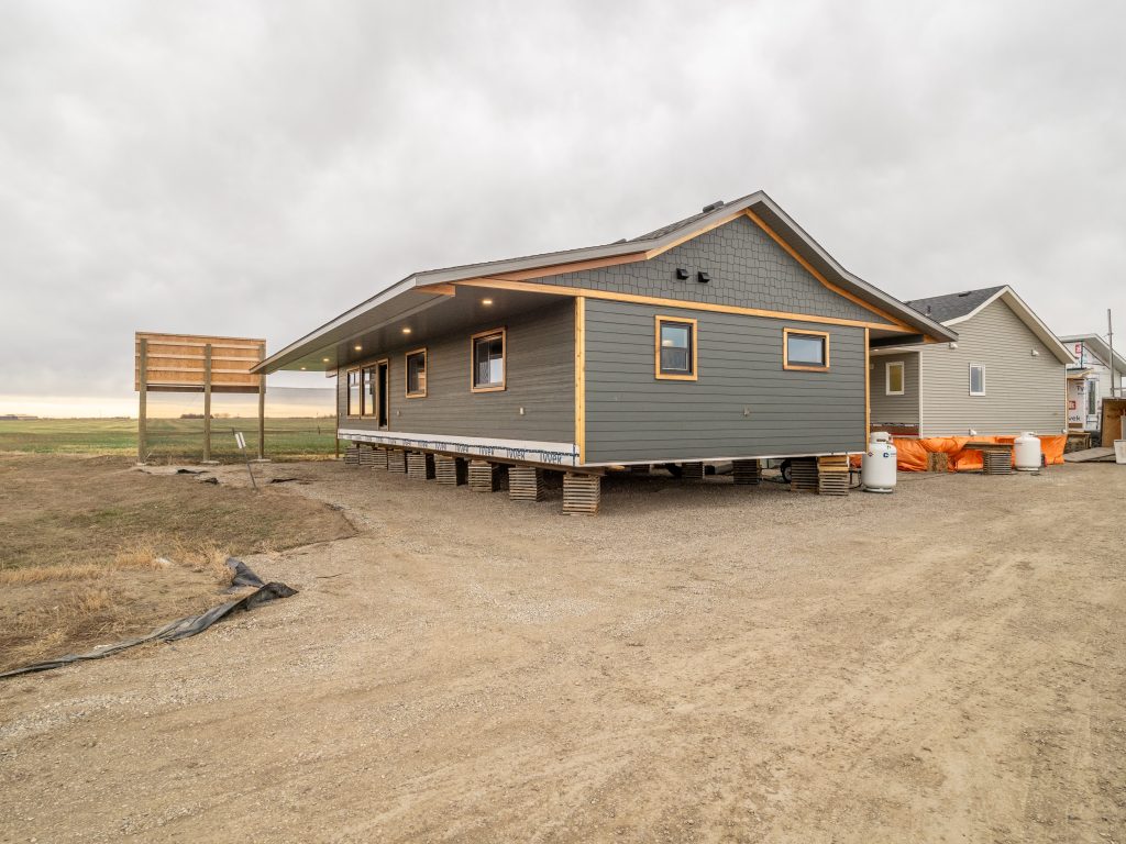 Single-story gray house on stilts, set on a gravel lot with sparse grass. Overcast sky, creating a calm, rural setting.