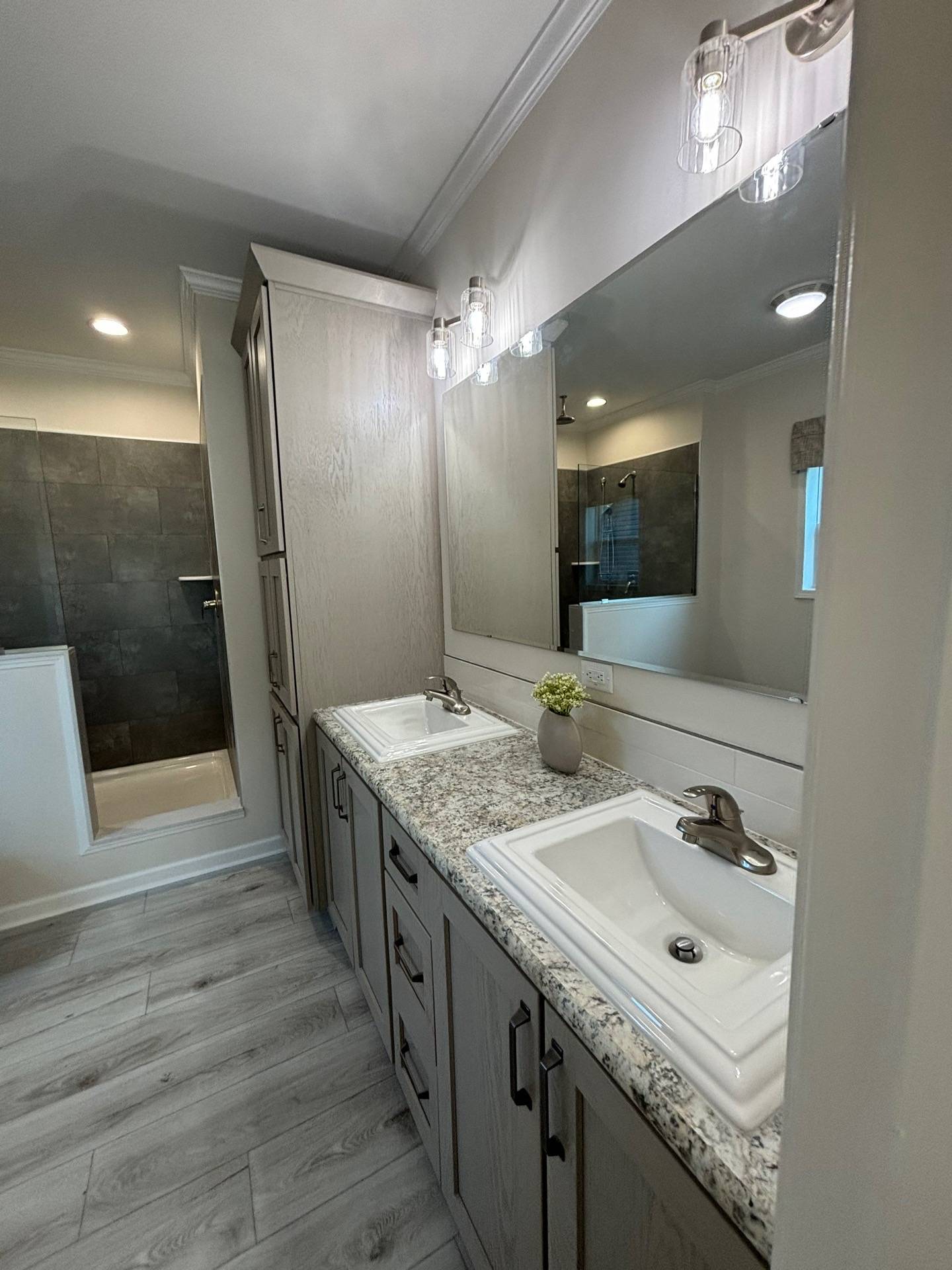Spacious bathroom featuring dual sinks with granite countertops, large mirror, gray cabinets, and a modern shower. Neutral tones create a calm vibe.