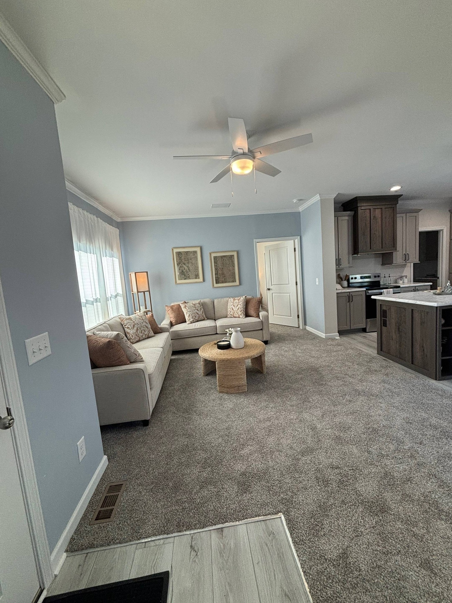 Spacious living room with light blue walls and grey carpet. Features a beige sectional sofa with pillows, wooden coffee table, and a ceiling fan.