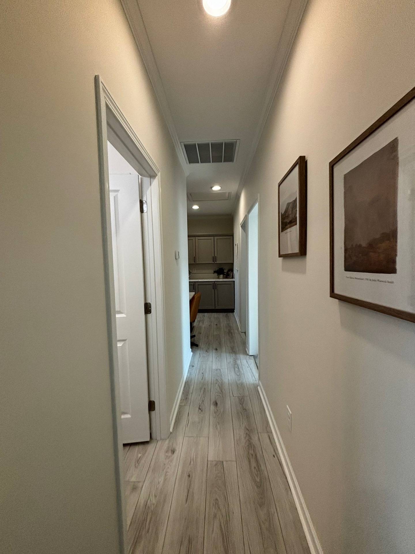 Narrow hallway with light wood flooring, leading to a kitchen. White walls feature framed artwork. A soft, warm light enhances a cozy atmosphere.