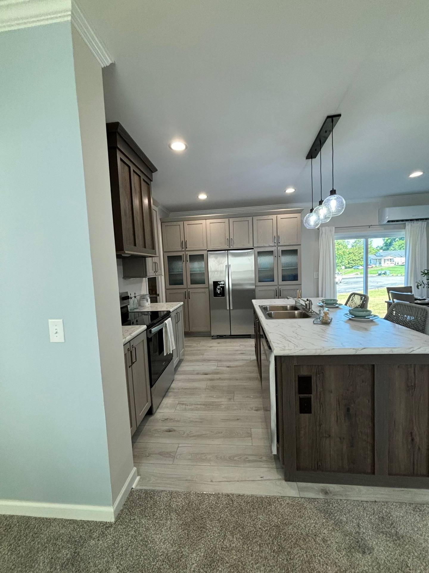 Modern kitchen with wooden cabinets, stainless steel appliances, and a marble island. Bright lighting, open layout, and a large window create a welcoming atmosphere.