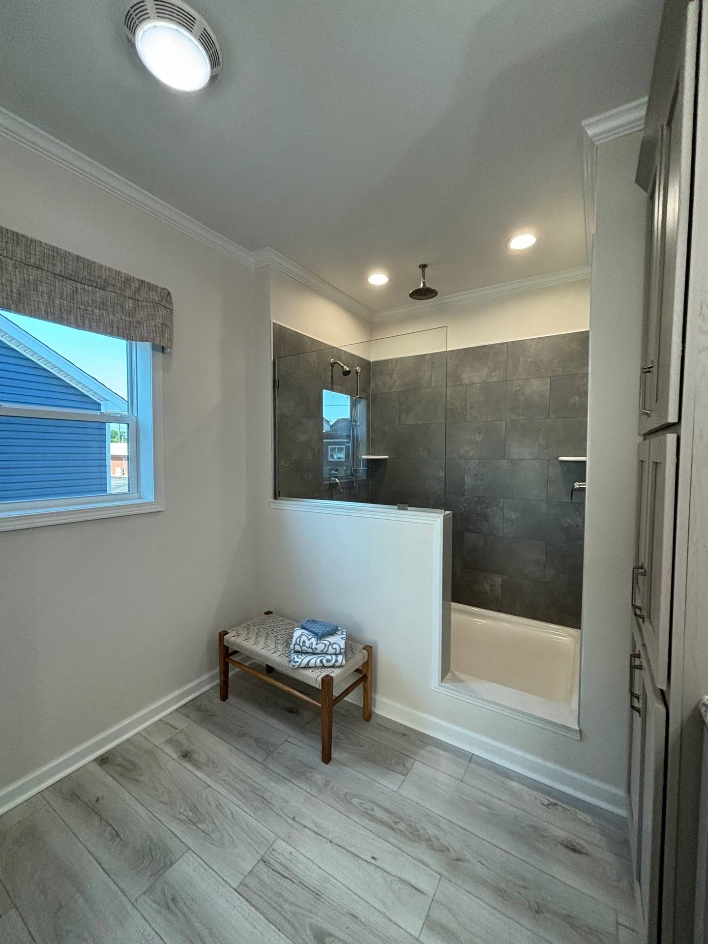 Modern bathroom with a glass-walled shower featuring dark tiles and an overhead rain showerhead. A wooden bench with folded towels sits nearby.
