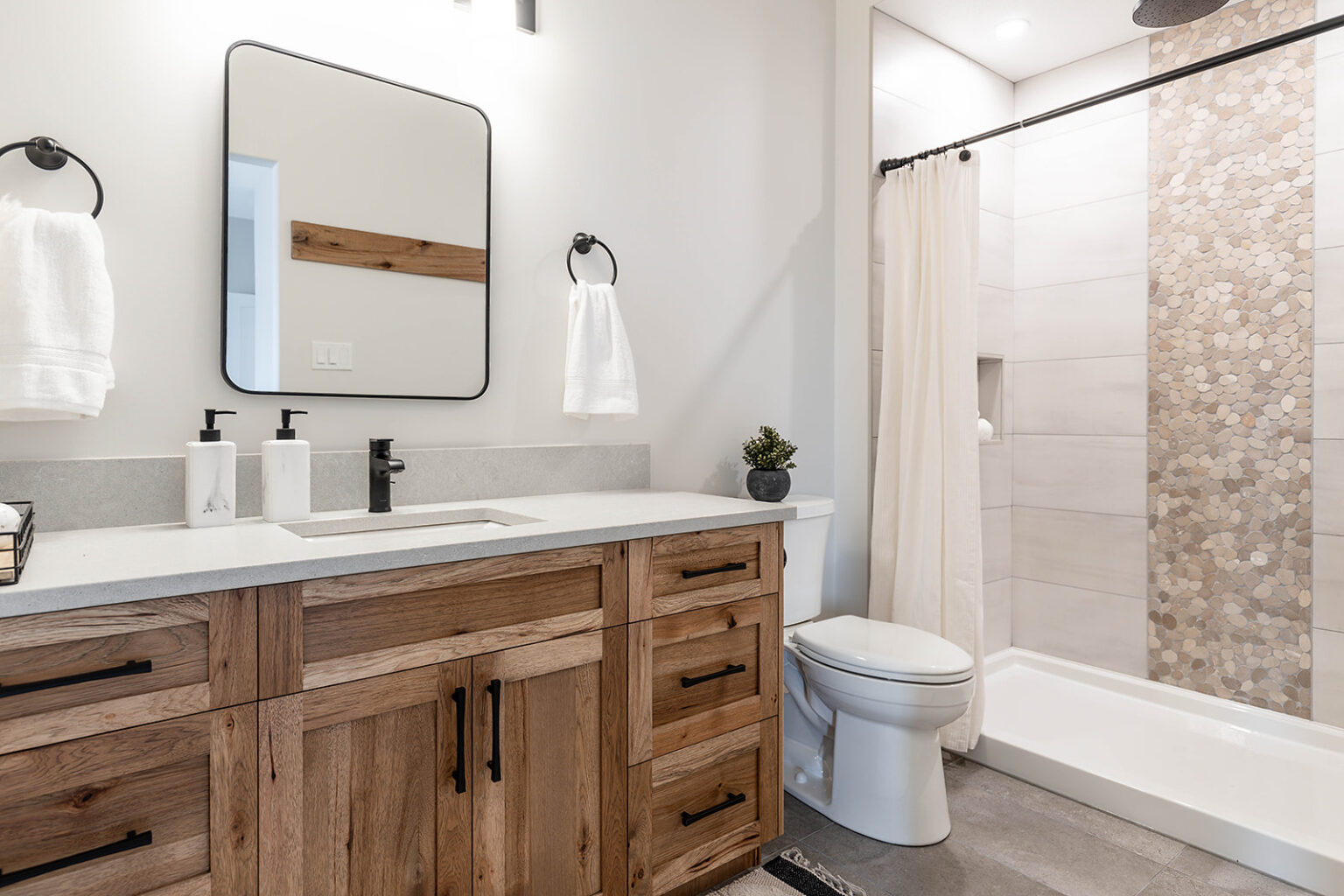 Modern bathroom with a wooden vanity, black hardware, and a large mirror. Features a white countertop, towels, a toilet, and a shower with tile accents. Cozy and clean.