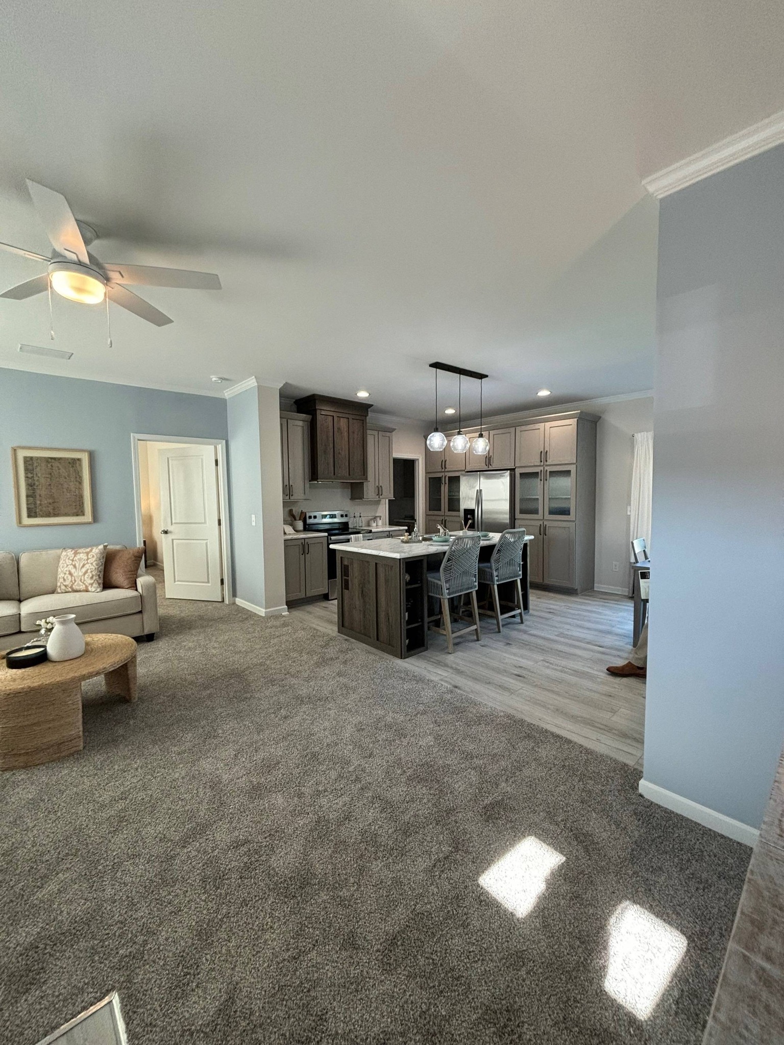 Modern open-concept living space with gray carpet, a cozy beige sofa, and a light-filled kitchen featuring an island with four barstools.