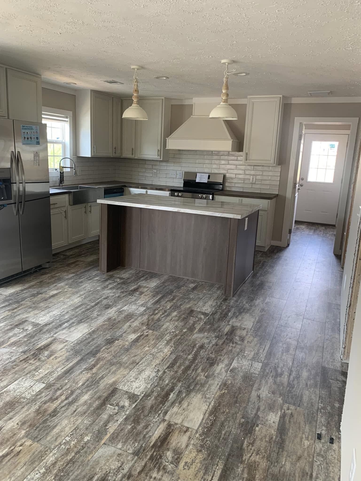 Modern kitchen with wood-look flooring, light gray cabinets, and a central island. Stainless steel appliances and white subway tile backsplash. Bright and inviting.