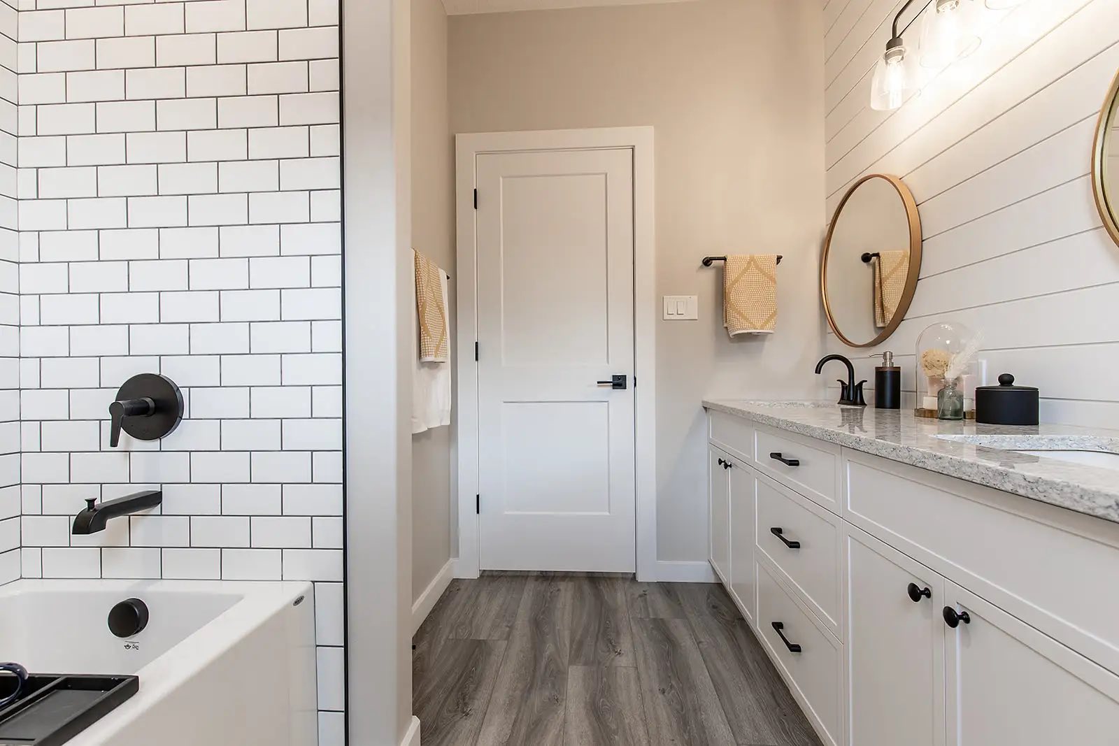 Chic bathroom with white subway tiles, black fixtures, dual round mirrors, shiplap walls, gray wood flooring, and soft lighting creating a cozy ambiance.