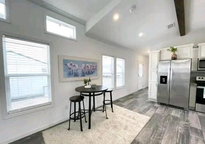 Modern kitchen with white walls, wood floors, stainless steel fridge, and a round table with two stools on a rug. Bright and airy atmosphere.