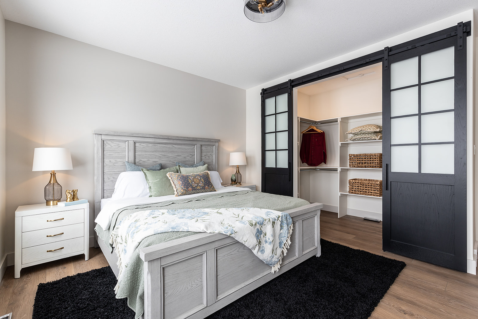 A cozy bedroom features a gray wooden bed with floral bedding, flanked by two white nightstands and lamps. An open closet with sliding doors reveals organized shelves and hanging clothes, creating a tidy and inviting atmosphere.