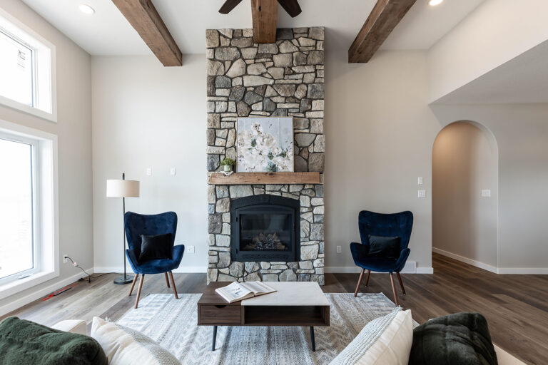 A cozy living room with a stone fireplace and wood beam ceiling. Two blue chairs flank the fireplace, exuding warmth and modern elegance.