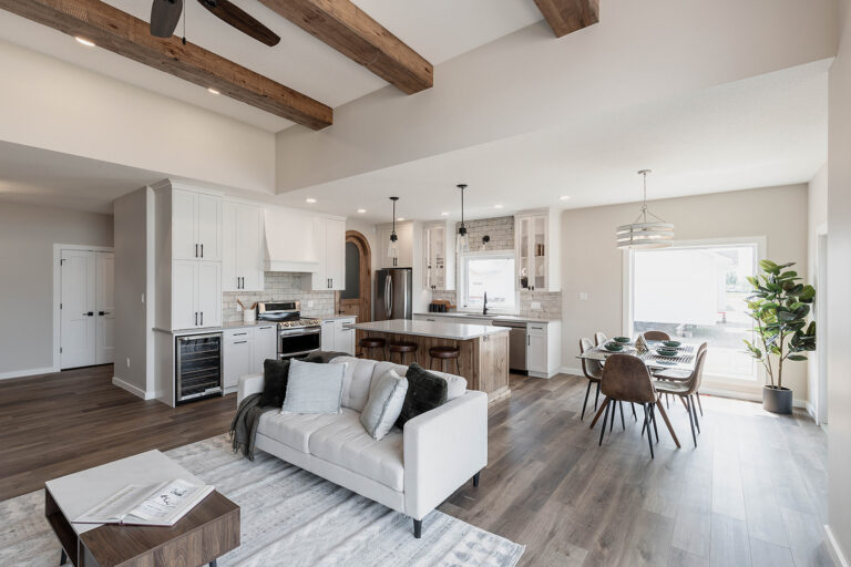 Spacious open-plan living area with wooden beams, a modern kitchen, island, cozy sofa, dining table by large window, and a calming neutral palette.