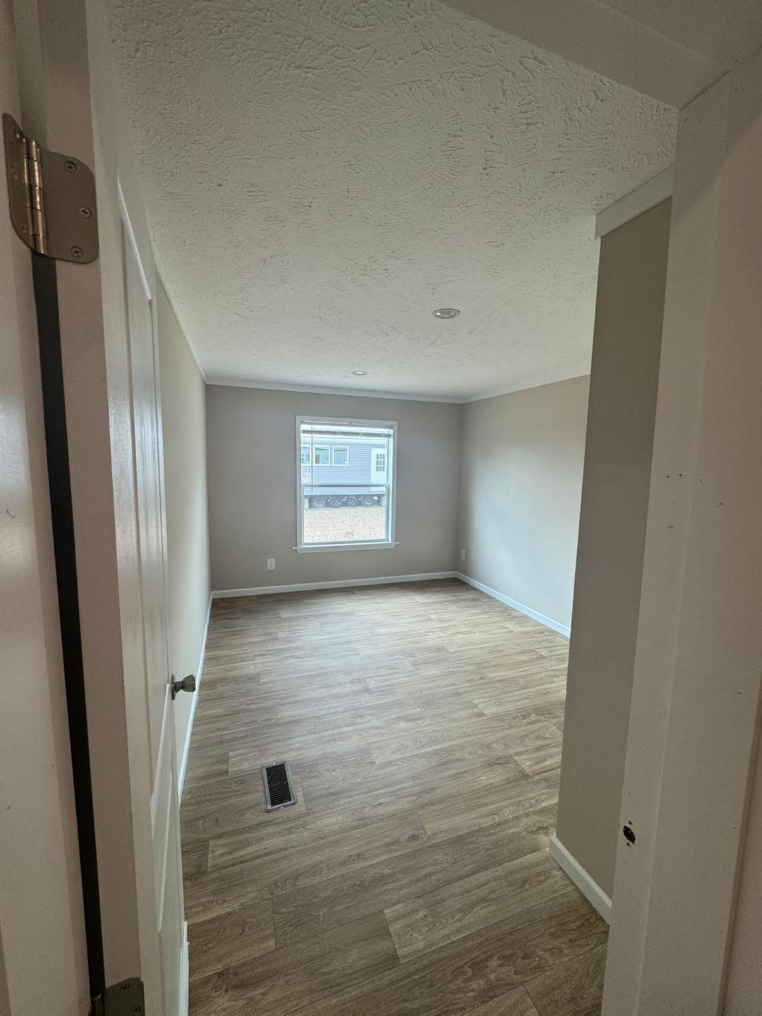 Empty room with light wood flooring and cream walls, viewed from an open door. A window at the far end lets in natural light, creating a calm atmosphere.