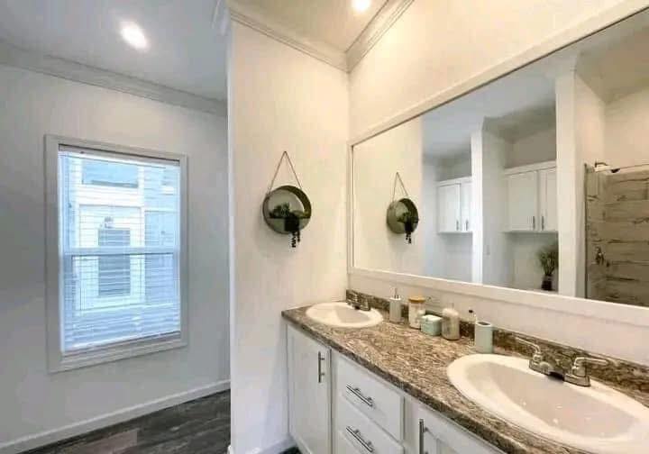Modern bathroom with dual sinks on a granite countertop, large mirror, and circular wall decor. Bright lighting and a window enhance the clean, spacious feel.