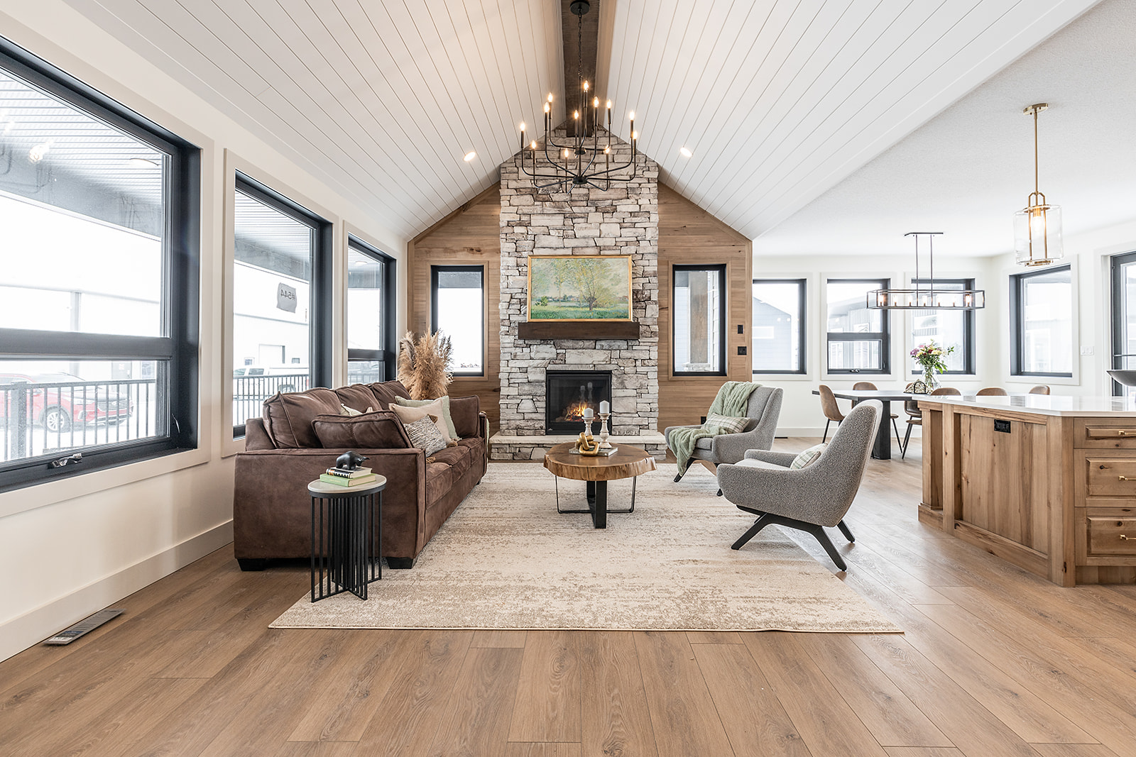 Spacious living room with vaulted ceilings, stone fireplace, and modern chandelier. Features a brown leather sofa, gray chairs, and large windows. Cozy and inviting atmosphere.