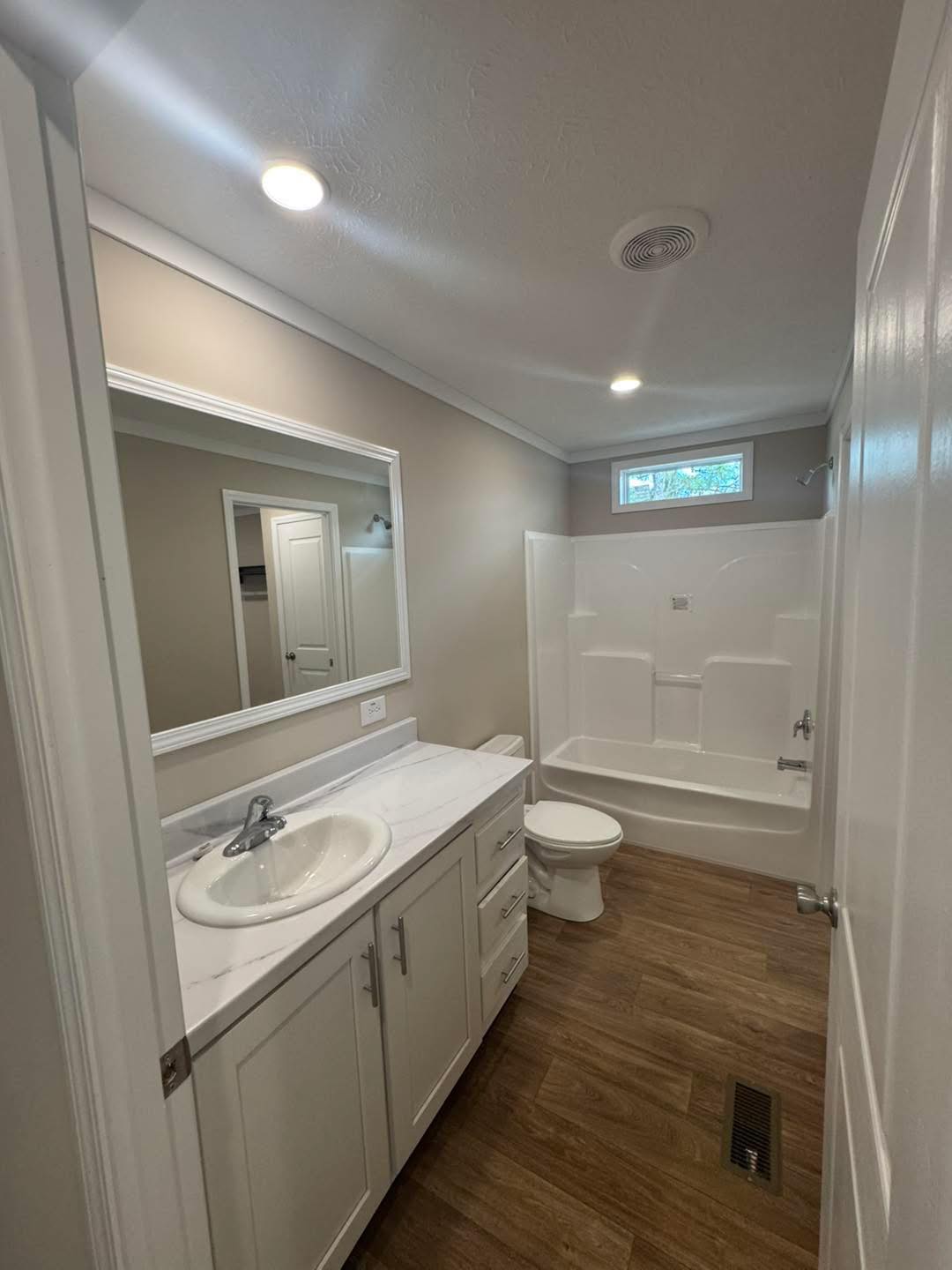 Bright bathroom with wood floors, white vanity with sink, large mirror, and a shower-tub combo. Neutral tones create a clean, modern feel.