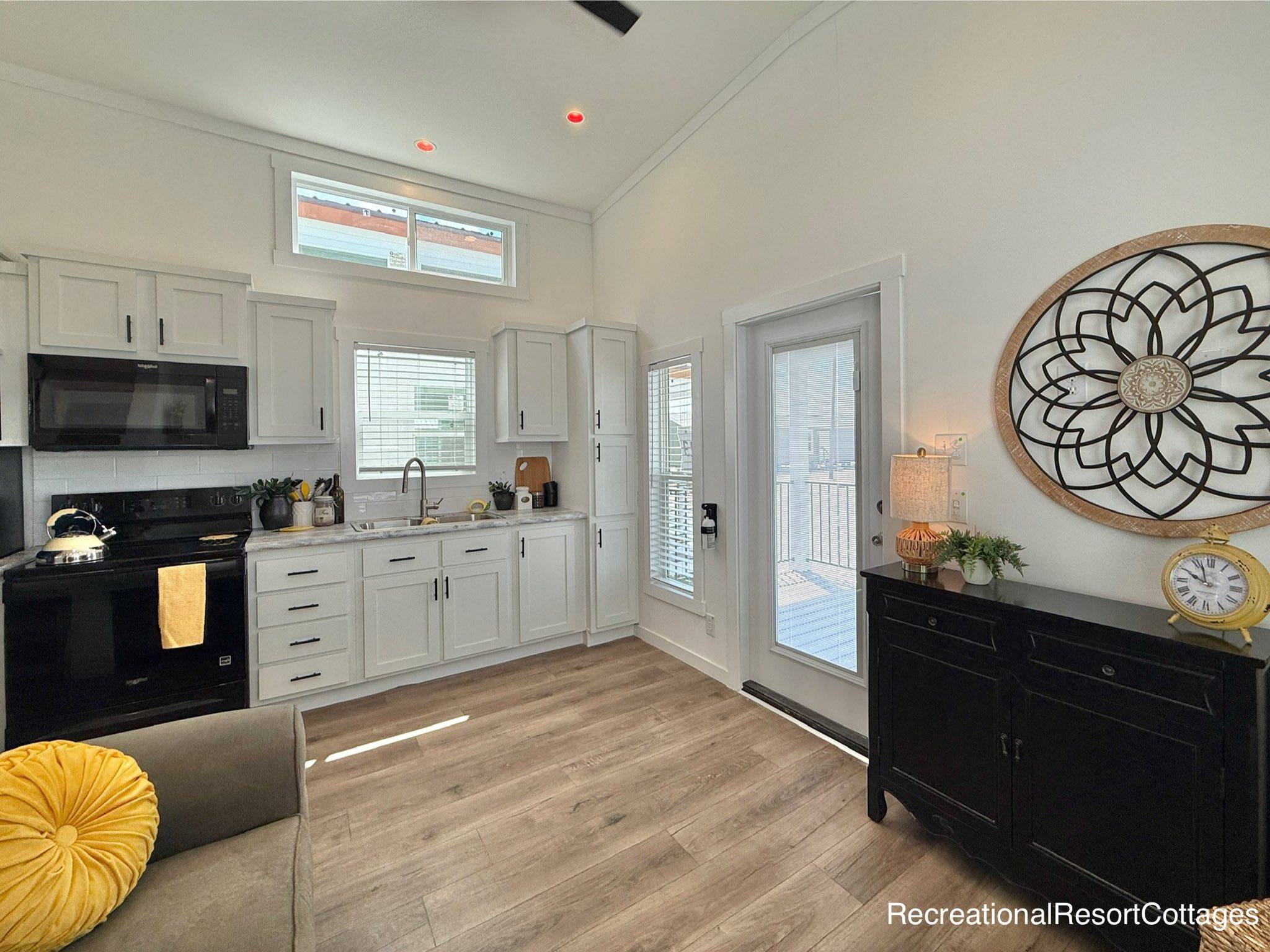Bright, modern kitchen with white cabinetry, black appliances, and wooden floors. A decorative clock and lamp enhance the cozy atmosphere.