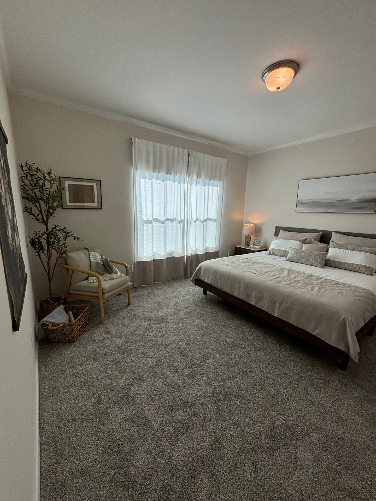 A serene bedroom with a comfortable double bed, soft gray carpet, sheer curtains, and gentle lighting. A cozy chair and wall art add to the peaceful ambiance.
