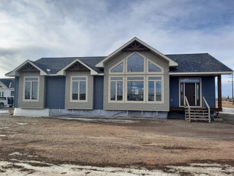 A newly constructed blue house with large front windows and a gabled roof sits against a cloudy sky. The ground is bare dirt with patches of snow.
