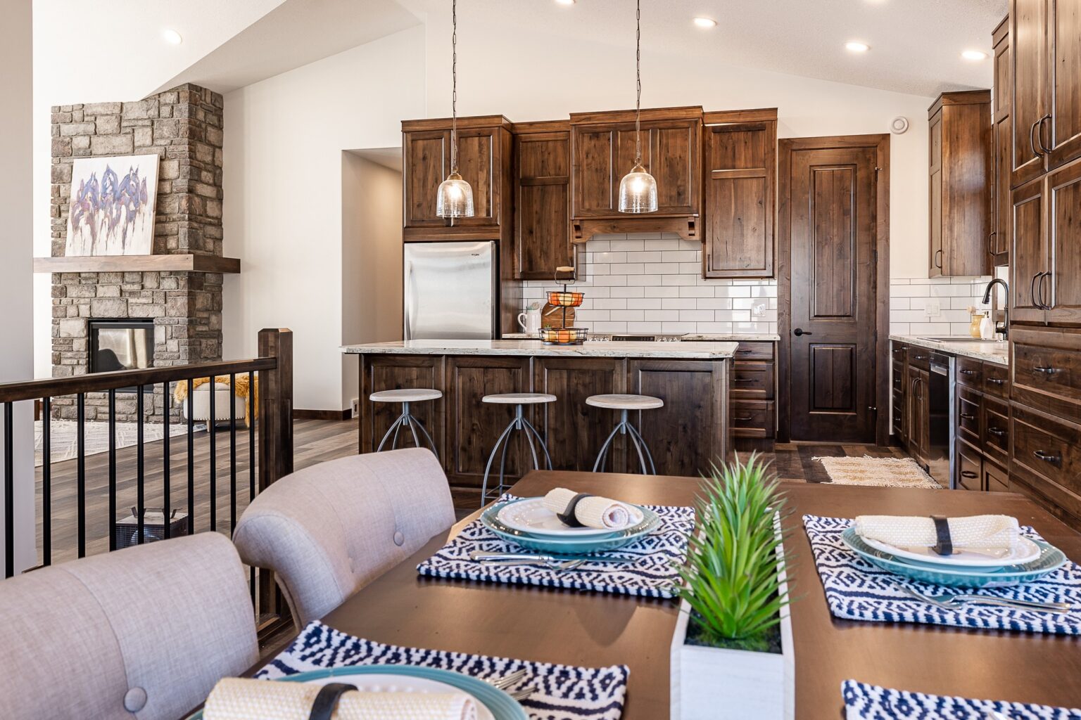 Warm, rustic kitchen with dark wood cabinets, white subway tiles, and a stone fireplace. A dining table with patterned placemats and a plant adds coziness.