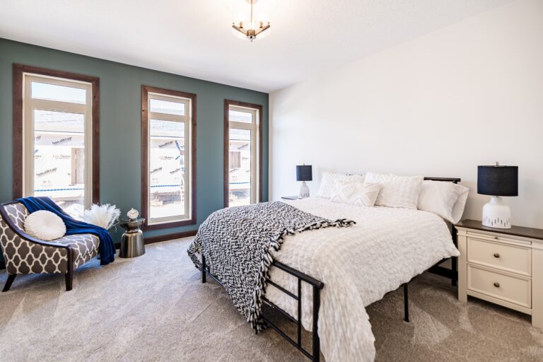 Bright bedroom with a cozy white bed, patterned chair, and three tall windows. Soft light, neutral tones, modern décor evoke a peaceful ambiance.