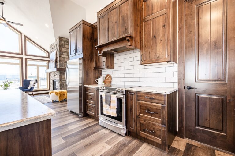 Bright kitchen with rustic wooden cabinets, white subway tile backsplash, and stainless steel appliances. Cozy living area visible in the background.