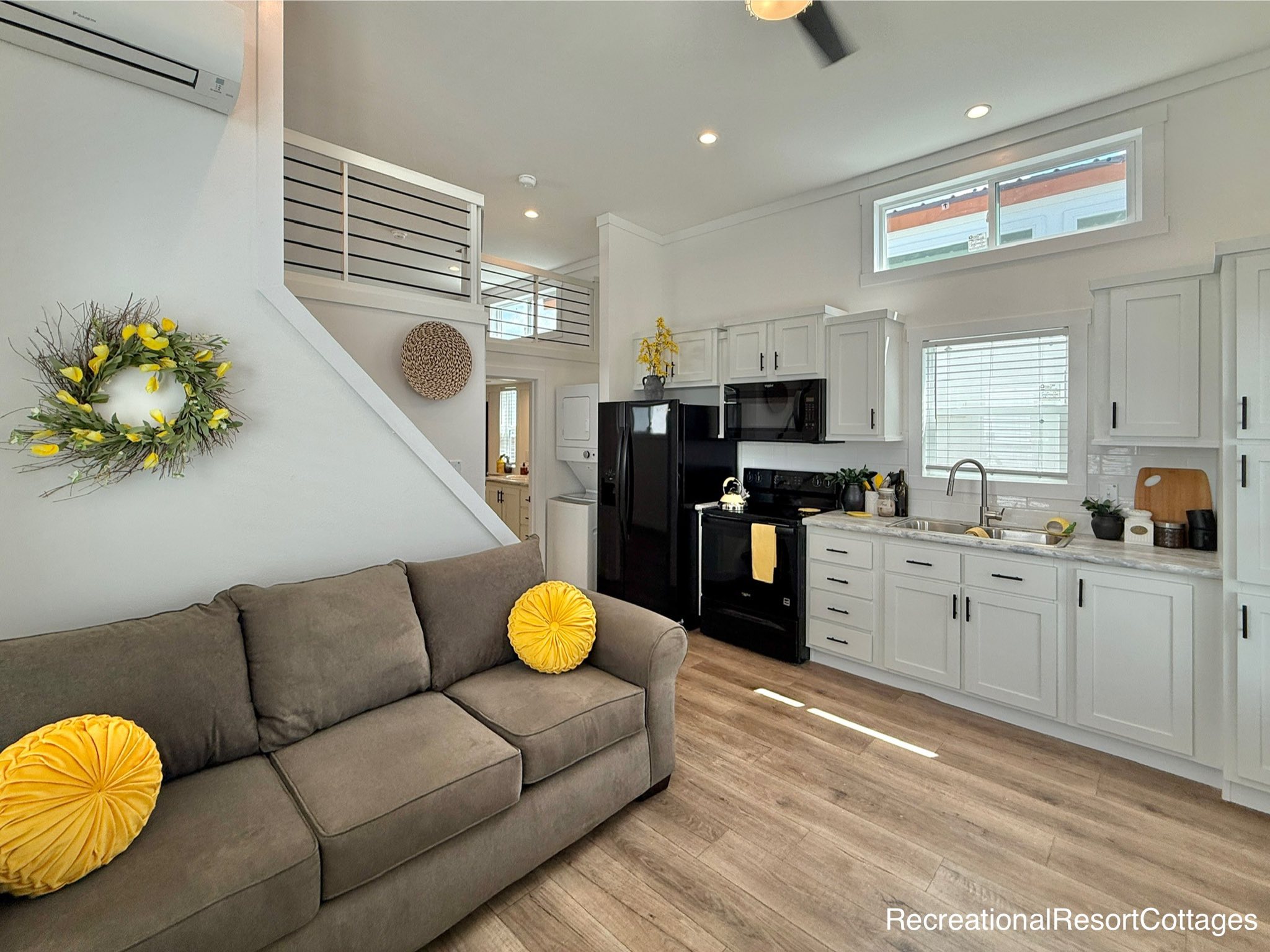 A modern, cozy living area with a gray sofa adorned with yellow pillows, a compact kitchen with sleek black appliances, and a loft space above.