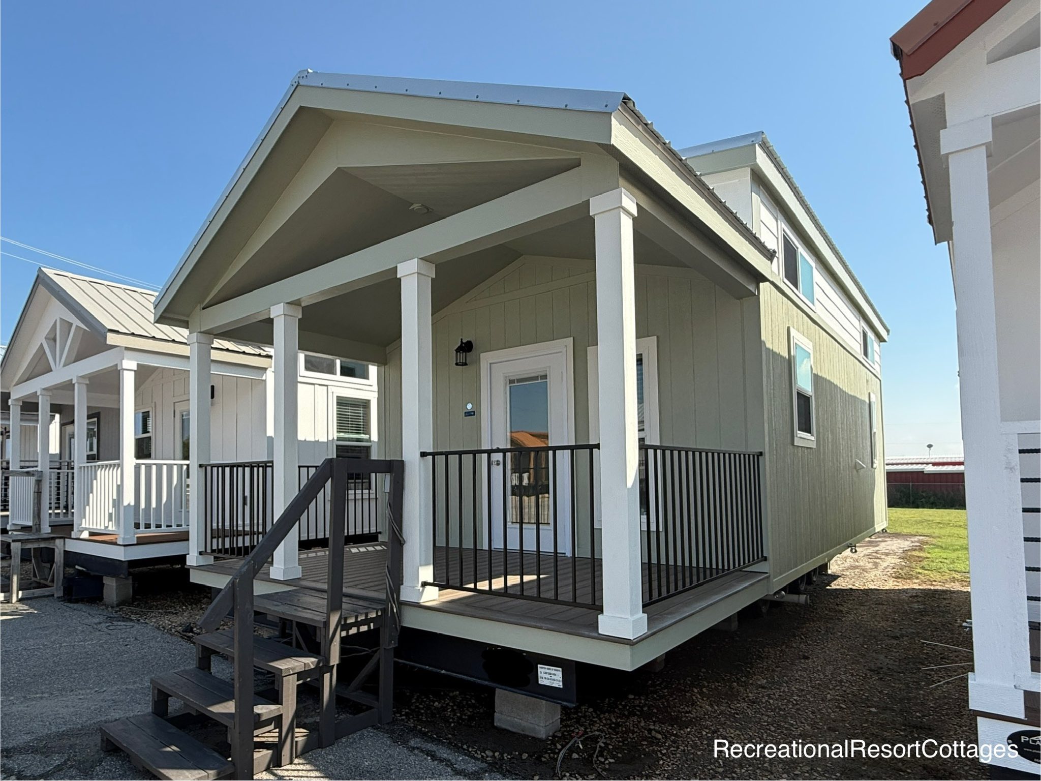 A modern tiny house with a green exterior and white trim, featuring a small front porch and steps. The sky is clear and sunny, conveying a tranquil atmosphere.